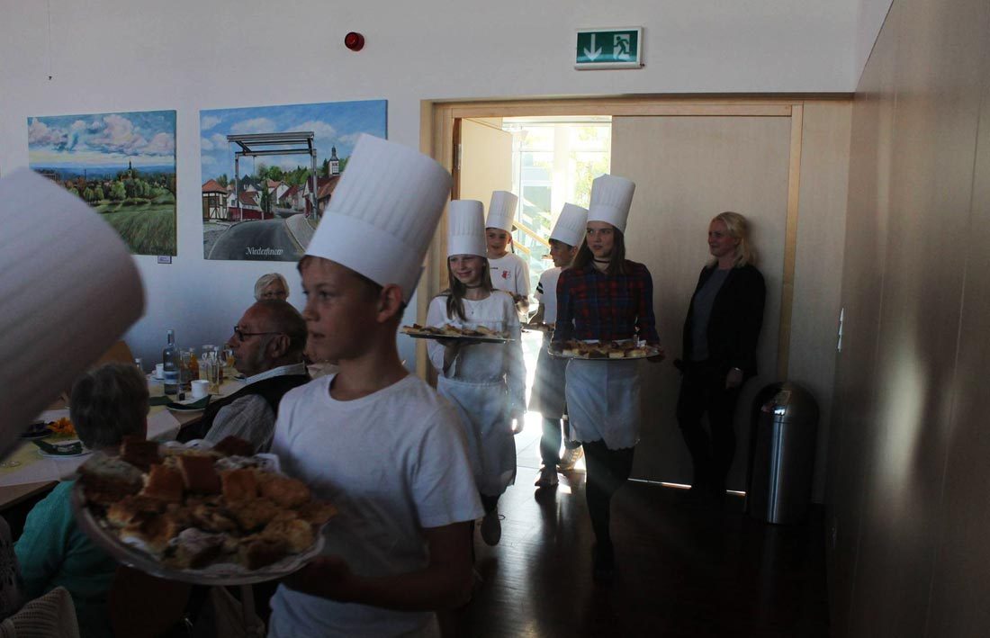 Kinder mit Bäckermützen tragen Kuchen in den Saal.