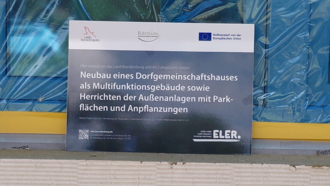 Schild: "Hier investieren das Land Brandenburg und die Europäische Union: Neubau eines Dorfgemeinschaftshauses als Multifunktionsgebäude sowie Herrichten der Außenanlagen mit Parkflächen und Anpflanzungen. ELER."