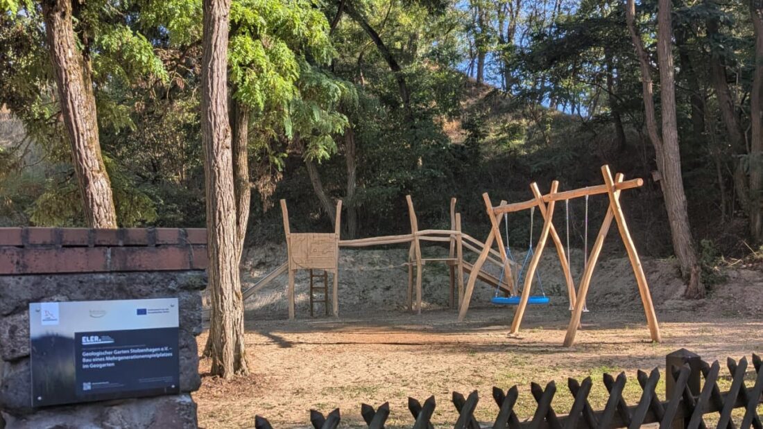 Spielplatz mit Klettergerüst und Schaukel in einem Waldgebiet. Im Vordergrund: Infotafel zur Förderung des Geologischen Gartens Stolzenhagen e. V. durch die EU und das Land Brandenburg.