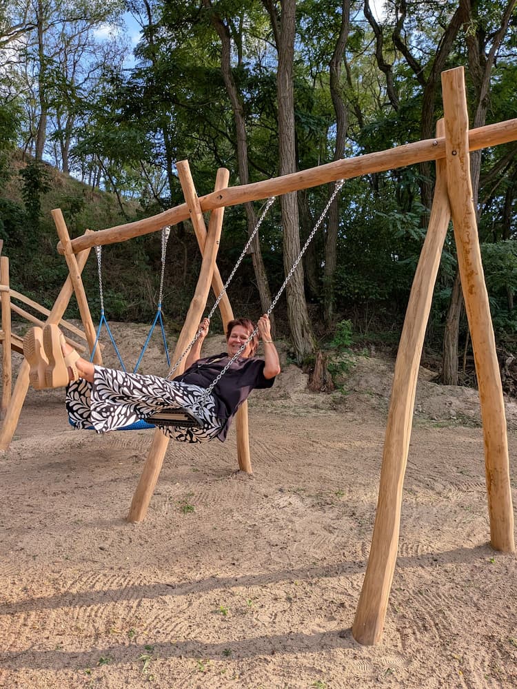 Die Bürgermeisterin der Gemeinde Lunow-Stolzenhagen schaukelt auf einer Holzschaukel, die zwischen zwei stabilen Holzpfosten aufgehängt ist, in einem sandigen Bereich mit Baumbestand im Hintergrund.