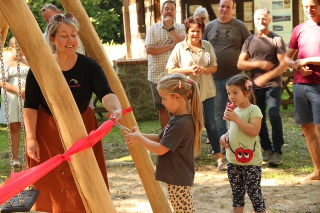 Feierliche Eröffnung eines Spielplatzes: Ein Kind durchschneidet mit einer Schere ein rotes Band, das zwischen zwei Holzpfosten gespannt ist. Eine Frau hält das Band, während eine Gruppe von Zuschauern im Hintergrund die Szene beobachtet.