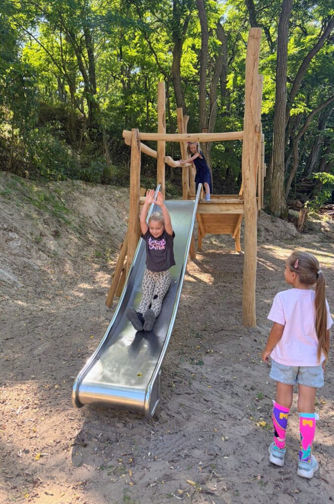 Drei Kinder auf einem Spielplatz: Ein Kind rutscht eine Edelstahlrutsche hinab, während ein weiteres Kind daneben steht. Die Rutsche ist in ein Holzgerüst integriert und von Bäumen umgeben.