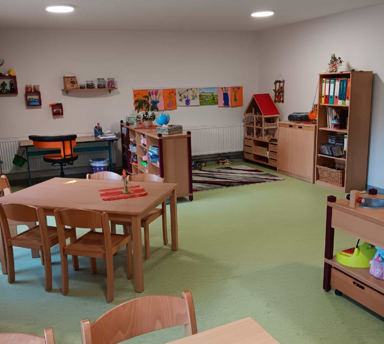 Gesamtansicht eines Kita-Gruppenraumes mit hellen Möbeln (Tische, Stühle, Schränke) und selbstgemalten Bildern an der Wand.